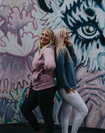 Two mums posing side on in front of a mural one wearing a dusty pinky breastfeeding hoodie